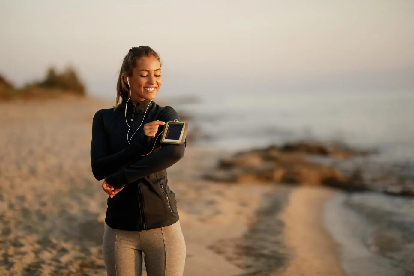 Blije sportieve vrouw bereidt zich voor op het hardlopen en past de muziek aan op haar smartphone op het strand.